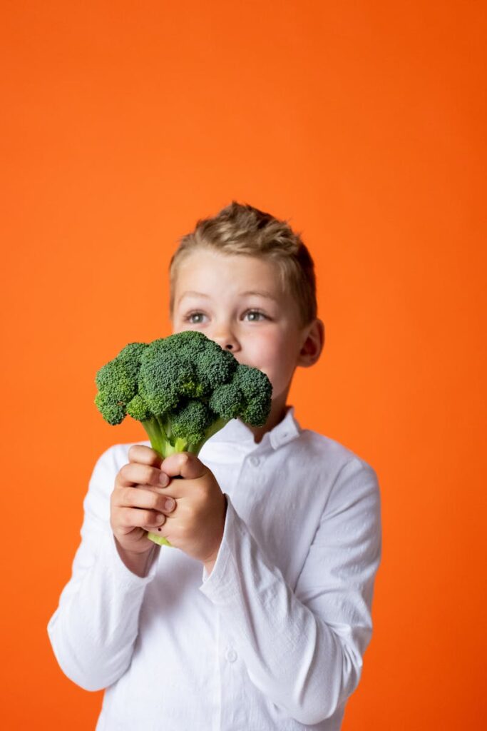 A cheerful young boy holds fresh broccoli, promoting healthy eating against a vibrant orange backdrop.