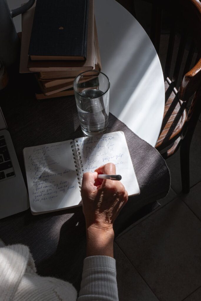 Hand writing in a notebook with books and a glass of water on a table, sunlight casting shadows.