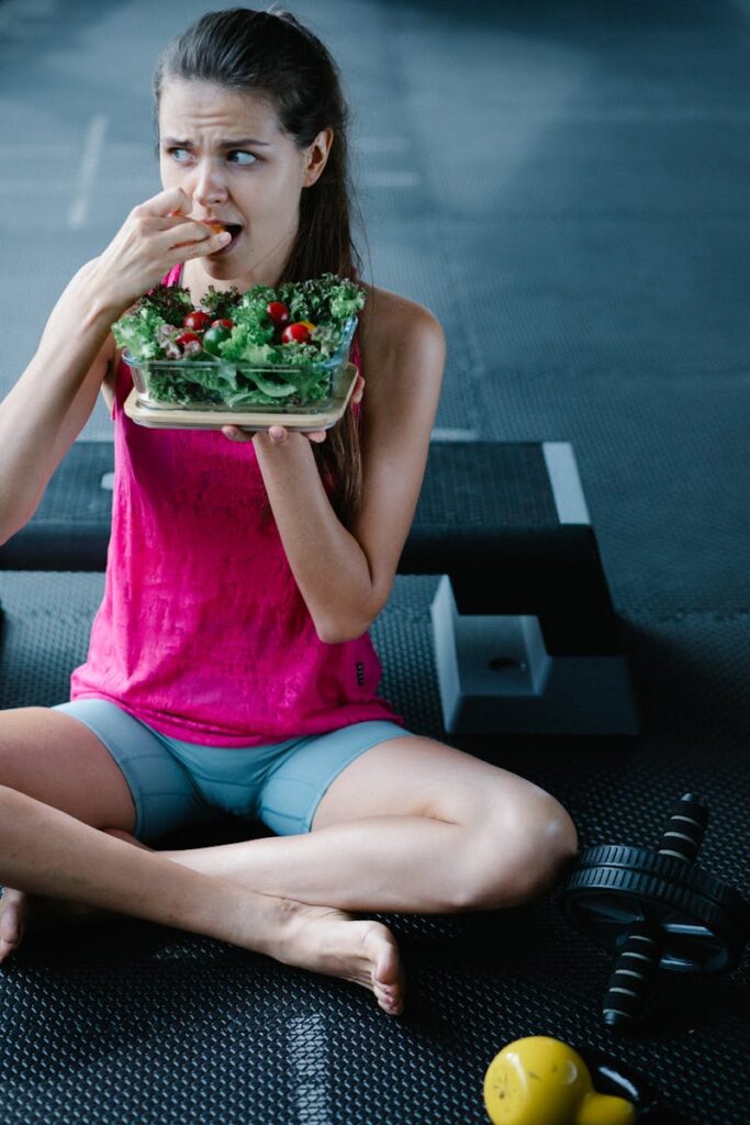 Woman in fitness clothing eats salad in a gym setting, promoting healthy lifestyle.